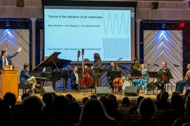Musicians perform a concert while a slide displays in the background explaining the science of sound.