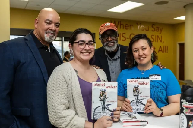 Two students hold copies of the Planetwalker book while standing with John Francis and Christopher Kilgour.